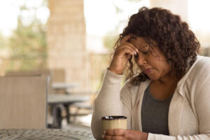 Woman with curly hair holding her forehead in one hand and a mug in the other, wearing a light sweater, seated outdoors with a blurred patio background. is emotional distress a personal injury